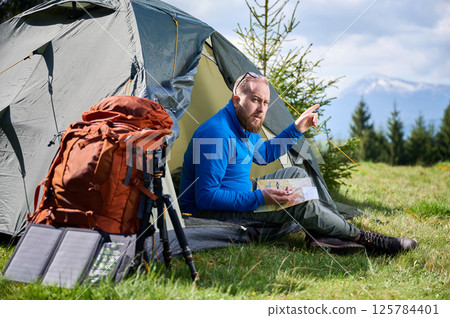 Man sits in tourist tent's entrance, points into distance while studying map. Beside him orange backpack, equipped with tripod and solar panel, set against scenic background of trees and mountains. Man sits in tourist tent's entrance, points into distance while studying map. Beside him orange backpack, equipped with tripod and solar panel, set against scenic background of trees and mountains. 125784401