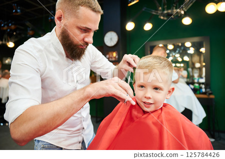 Man barber cutting little boy's hair using comb and scissors. Child getting haircut from adult male, likely barber. Professional hairdresser and cute client at modern barbershop. 125784426