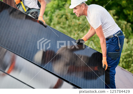 Workers building solar panel system on roof of house. Two men installers in helmets carrying photovoltaic solar module outdoors. Alternative, green and renewable energy generation concept. 125784429