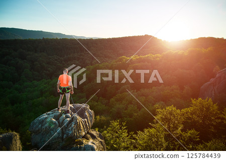 Aerial view of climber standing atop rock formation at Dovbush Rocks in Carpathian mountains, Ukraine. Sun sets, casting warm glow over lush, green forest, distant hills, creating breathtaking view. 125784439