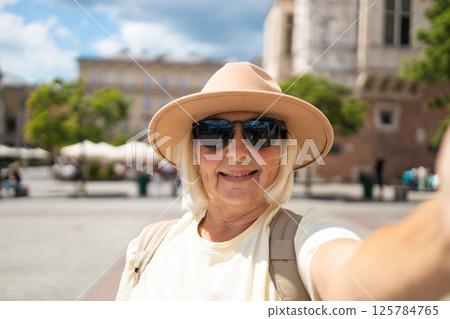 Senior woman taking a selfie, waving hand in a city square, enjoying vacation. 125784765