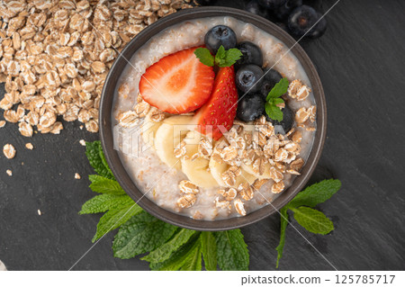 Cooked spelt in bowl. Oatmeal milk porridge with fruits and berries on dark background Cooked spelt in bowl. Oatmeal milk porridge with fruits and berries on dark background 125785717