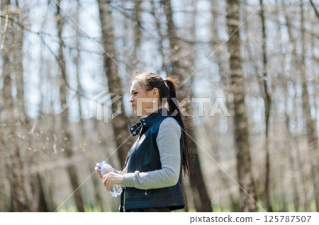 A beautiful girl in leather pants and a bomber jacket holds a bottle of water in a spring forest and basks in the sun. A young woman is walking in the park with a bottle of mineral water A beautiful girl in leather pants and a bomber jacket holds a bottle of water in a spring forest and basks in the sun. A young woman is walking in the park with a bottle of mineral water 125787507