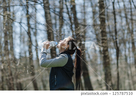A young woman drinking water in the forest. A woman resting in a park and drinking mineral water from a plastic bottle. 125787510