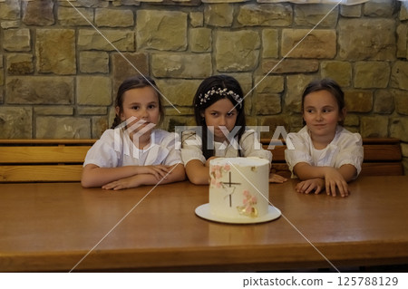 The girls in matching blouses and a dress form a charming group around the cake. The twin girls with two braids look fascinated. The girls in matching blouses and a dress form a charming group around the cake. The twin girls with two braids look fascinated. 125788129