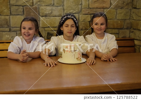 The cake decorates the center of the table, surrounded by joyful girls. The older sister and the twins admire each element of the dessert with fascination. The cake decorates the center of the table, surrounded by joyful girls. The older sister and the twins admire each element of the dessert with fascination. 125788522