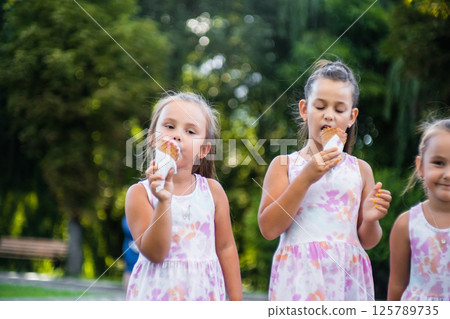Three sisters stand side by side in a park enjoying an ice cream. 125789735