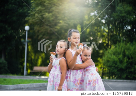 Three sisters, dressed in identical dresses, stand holding hands and enjoying ice cream. Three sisters, dressed in identical dresses, stand holding hands and enjoying ice cream. 125789812