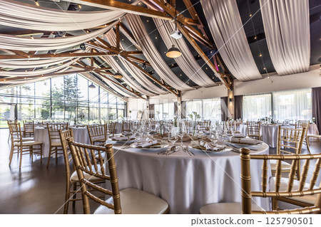 Wedding setting and decorated tables in a beautiful modern interior of a performance hall with a modern light ceiling of wooden construction Wedding setting and decorated tables in a beautiful modern interior of a performance hall with a modern light ceiling of wooden construction 125790501