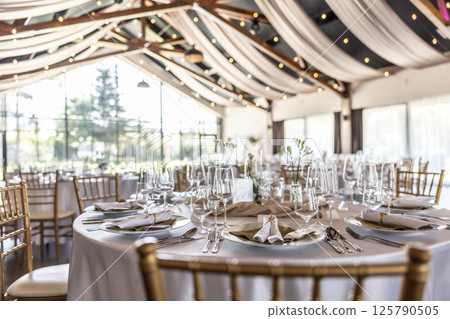 Wedding setting and decorated tables in a beautiful modern interior of a performance hall with a modern light ceiling of wooden construction Wedding setting and decorated tables in a beautiful modern interior of a performance hall with a modern light ceiling of wooden construction 125790505