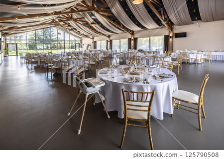 Wedding setting and decorated tables in a beautiful modern interior of a performance hall with a modern light ceiling of wooden construction Wedding setting and decorated tables in a beautiful modern interior of a performance hall with a modern light ceiling of wooden construction 125790508