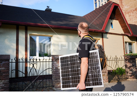 Man holding photovoltaic solar panel in front of historical building. Concept of integration of sustainable renewable energy sources into architecture. Man holding photovoltaic solar panel in front of historical building. Concept of integration of sustainable renewable energy sources into architecture. 125790574