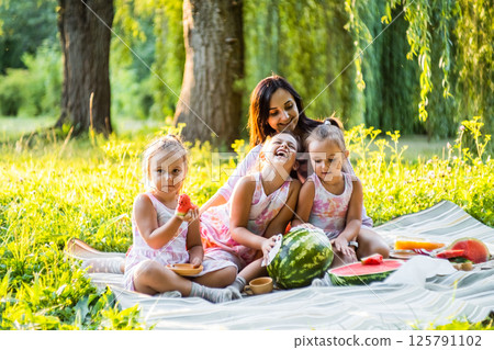 The girls and their mother are eating watermelon, their dresses are a little dirty. The twins do not pay attention, laughing together.  125791102