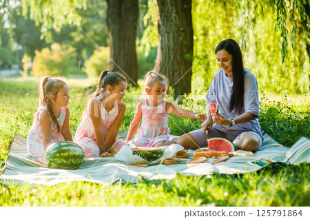 Cute little girls of 4-5 years old and their mother are eating a delicious watermelon against the background of green nature. Healthy lifestyle. Cute little girls of 4-5 years old and their mother are eating a delicious watermelon against the background of green nature. Healthy lifestyle. 125791864