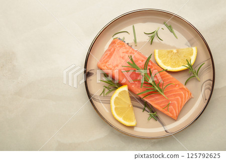 Red fish. Raw salmon fillet with rosemary and lemon on beige plate on grey background. 125792625
