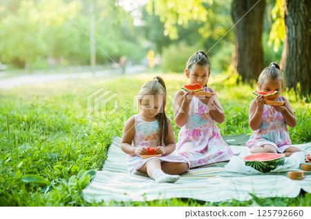 Three girls are holding pieces of watermelon in their hands. The beautiful girls are taking small bites of watermelon at a picnic. 125792660