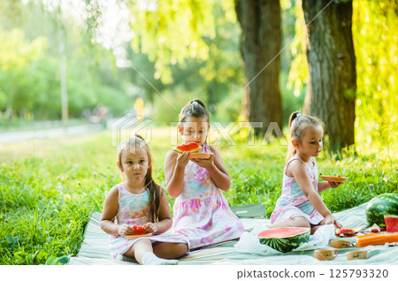 The sisters are sitting close to each other on a blanket. The little girls are holding pieces of watermelon in their hands. 125793320
