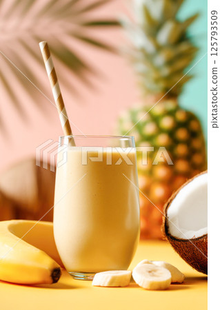 Tropical banana coconut pineapple smoothie in glass with striped straw on colorful summer background. 125793509