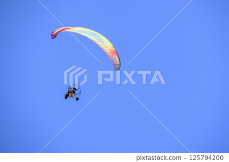 Paramotor glider with colorful parachute flying through a bright blue sky 125794200