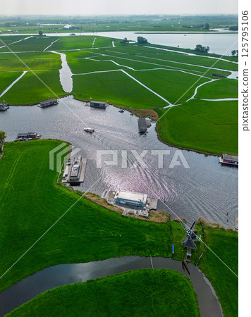 Aerial view of a peaceful Dutch landscape with green fields, a canal lined with houseboats, a traditional windmill, and a marina in the distance under a cloudy sky. Aerial view of a peaceful Dutch landscape with green fields, a canal lined with houseboats, a traditional windmill, and a marina in the distance under a cloudy sky. 125795106