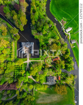 Aerial view of a historic castle surrounded by a forest, with vast green fields, winding waterways, and a river stretching across the flat Dutch countryside under a partly cloudy sky. Aerial view of a historic castle surrounded by a forest, with vast green fields, winding waterways, and a river stretching across the flat Dutch countryside under a partly cloudy sky. 125795112