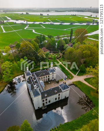 Aerial view of a historic castle surrounded by a forest, with vast green fields, winding waterways, and a river stretching across the flat Dutch countryside under a partly cloudy sky. Aerial view of a historic castle surrounded by a forest, with vast green fields, winding waterways, and a river stretching across the flat Dutch countryside under a partly cloudy sky. 125795115