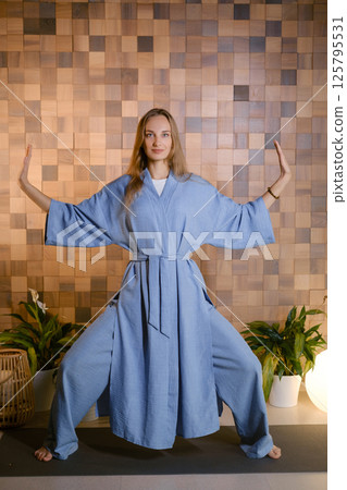 Yoga instructor in blue kimano at fitness gym . The girl does qigong yoga. The exercises are based on the movement of internal energy, the system of channels, energy centers, chakras Yoga instructor in blue kimano at fitness gym . The girl does qigong yoga. The exercises are based on the movement of internal energy, the system of channels, energy centers, chakras 125795531