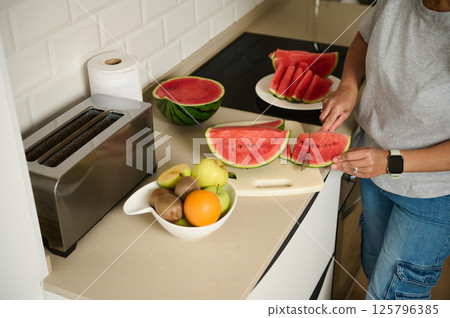 Person Slicing Fresh Watermelon in Kitchen with Fruits and Toaster 125796385
