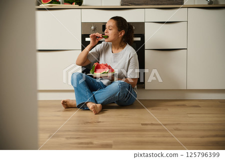 Woman Enjoying Slices of Watermelon in a Cozy Kitchen Setting 125796399