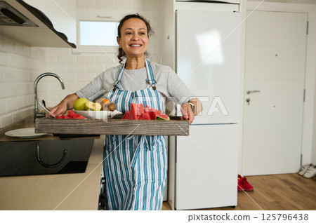 Smiling Woman Serving Fresh Fruit Tray in Bright Modern Kitchen 125796438