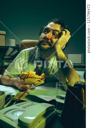 Daydreaming office worker holding sandwich, looks up with bored face at cluttered desk, showing fatigue 125796472