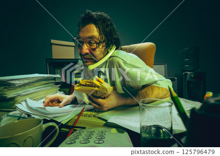 Man at office desk nervously checking documents, holding sandwich, talking on phone, showing stress, absurd, workload, burnout and multitask. 125796479