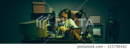 Man buried in documents, balances two phones while working under lamp in cluttered office, expressing burnout, fatigue, pressure. Man buried in documents, balances two phones while working under lamp in cluttered office, expressing burnout, fatigue, pressure. 125796482