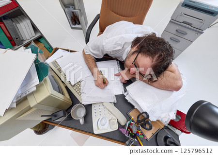 Top view of man, overworked office worker sitting at cluttered office desk, attempting to write. Extreme fatigue, burnout, large workload 125796512