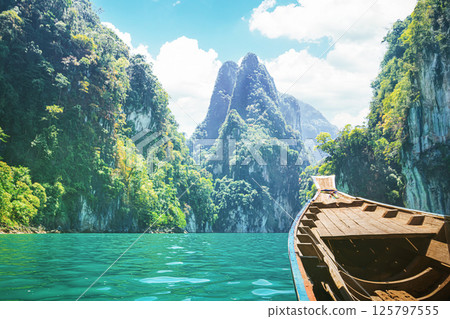 Travelers enjoy a cruise on a traditional longtail boat in Cheow Lan Lake, surrounded by towering limestone cliffs and lush greenery. The vibrant blue waters and scenic mountains 125797555