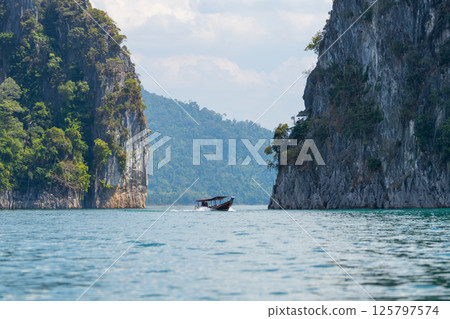 Small boat glides across the turquoise waters of Cheow Lan Lake, by towering limestone cliffs covered in lush greenery. Bright sunlight reflects on the lake's surface, enhancing the beauty of Khao Sok 125797574