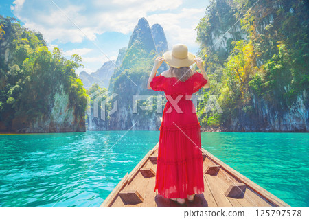 Woman female traveler enjoys the breathtaking views of Cheow Lan Lake, surrounded by towering limestone cliffs and lush greenery in Khao Sok, Thailand. Wearing a vibrant red dress and wide-brimmed hat 125797578