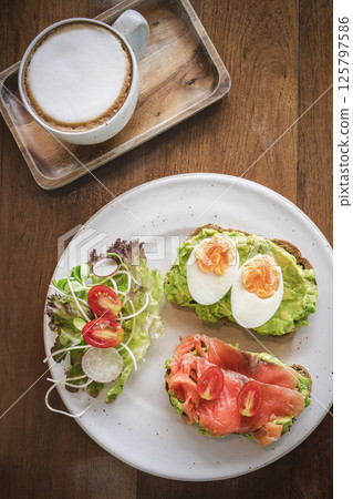 Delicious breakfast featuring avocado toast and smoked salmon with coffee on a wooden table, flatlay. Delicious breakfast featuring avocado toast and smoked salmon with coffee on a wooden table, flatlay. 125797586