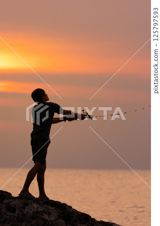 fisherman casting a spinning rod into the sea from a cliff against the background of an orange sea sunset, rear view close-up 125797593