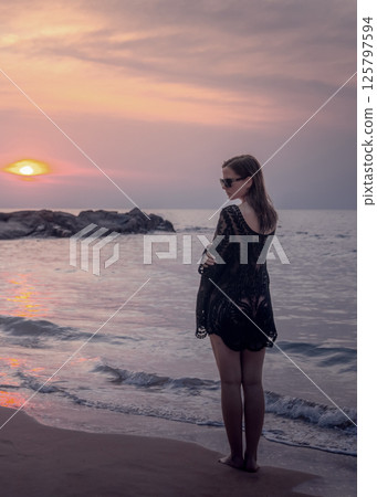 A woman in black beach clothes and sunglasses meets the sunset on the coast of Kaolak, pose - half-turned back. Beautiful view of the rocky seashore. Thoughtful reflection. 125797594