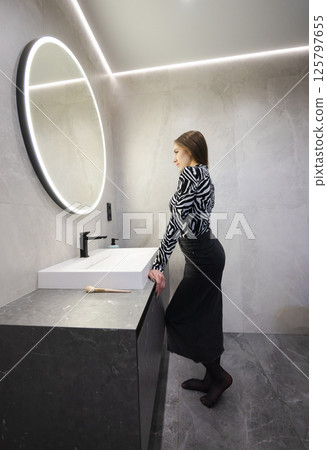 Teenage girl standing in a modern bathroom, leaning on the sink and looking at her reflection in a round illuminated mirror 125797655