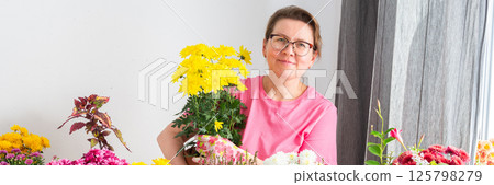 Woman 50 years old transplanting autumn flowers chrysanthemums and heathers into pots and holding a pot with a chrysanthemum and smiling, decorating a home terrace or balcony with flowers, banner Woman 50 years old transplanting autumn flowers chrysanthemums and heathers into pots and holding a pot with a chrysanthemum and smiling, decorating a home terrace or balcony with flowers, banner 125798279