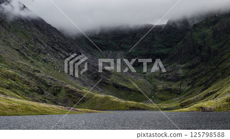 Coomloughra Lough lies serenely at the base of steep, green-clad mountains in Ireland. A blanket of clouds hovers above, enhancing the tranquil beauty of this natural setting. 125798588
