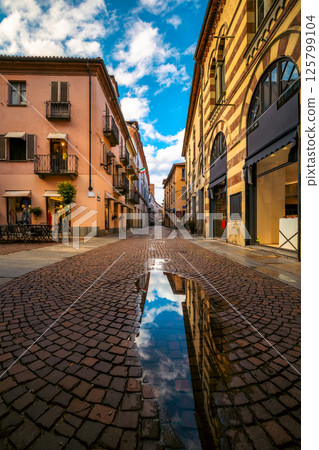 Cobblesone street among buildings under blue sky with white clouds in old town of Alba, Italy. 125799104