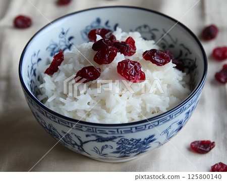 A bowl of white rice topped with dried cranberries sitting on a light colored tablecloth surface 125800140