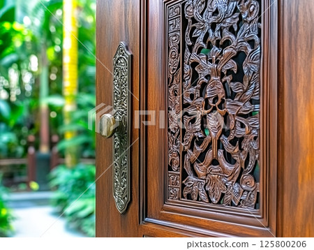 Close up of a carved wooden door with a brass handle and a garden view in the background outside 125800206