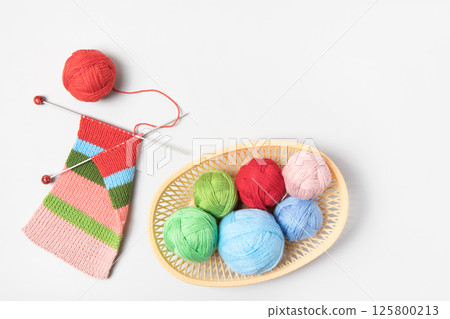 Red, green, blue and pink knitting balls in basket with knitting needles and knitted scarf on gray background. Set for the hobby of knitting warm clothes or toys. Concept needlework 125800213