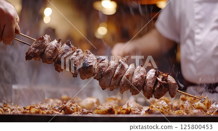 Close up of grilled meat on a skewer held by a person over a grill with other meat cooking on it 125800453