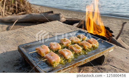 Salmon skewers with green sauce on a tray near a bonfire by the water on a sunny day outdoors 125800456