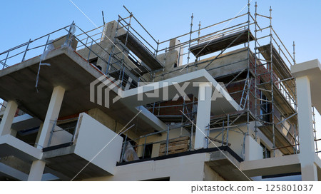 A house under construction is in its finishing stages, featuring scaffolding for workers. The structure shows modern design elements and reveals recent brickwork and concrete features. 125801037
